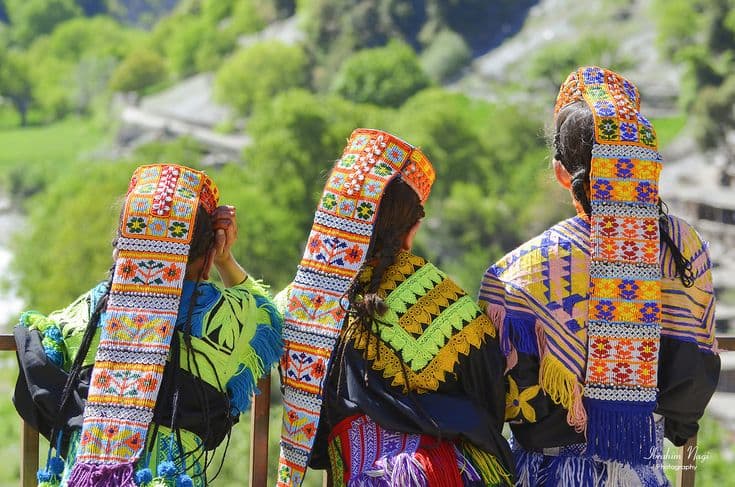 Three Kalash women in embroidered headdresses looking out over a Kalash valley